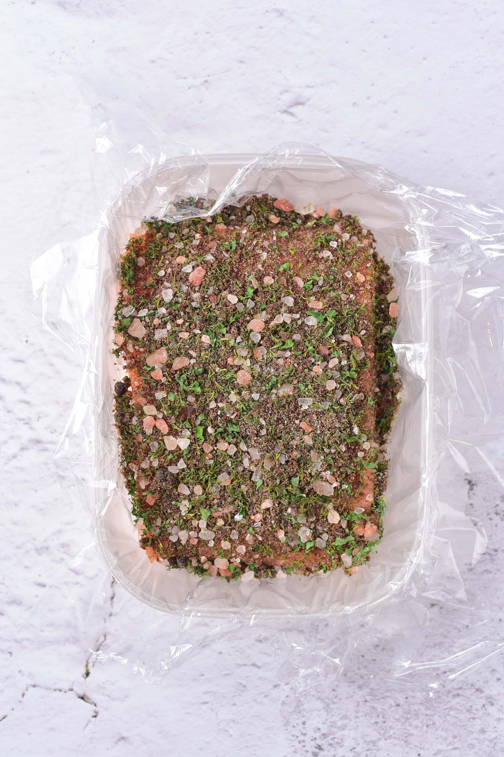 A piece of uncooked gravlax salmon covered with a mixture of dill, herbs, and coarse salt rests on a tray lined with plastic wrap. The background is a textured, light-colored surface. This easy recipe brings the classic Scandinavian dish right to your kitchen.