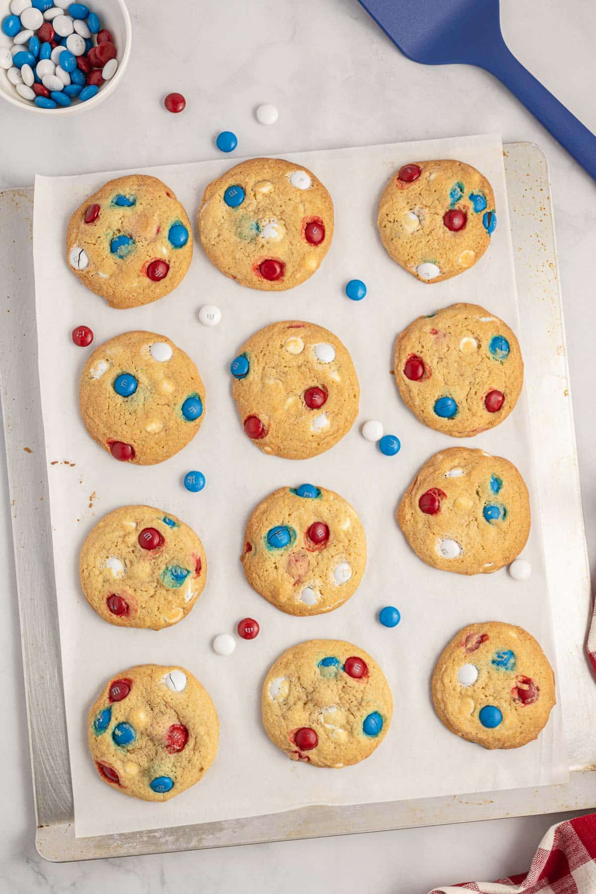 A baking sheet holds 12 cookies with red, white, and blue chocolate candies. A bowl of extra candy and blue spatula are nearby.