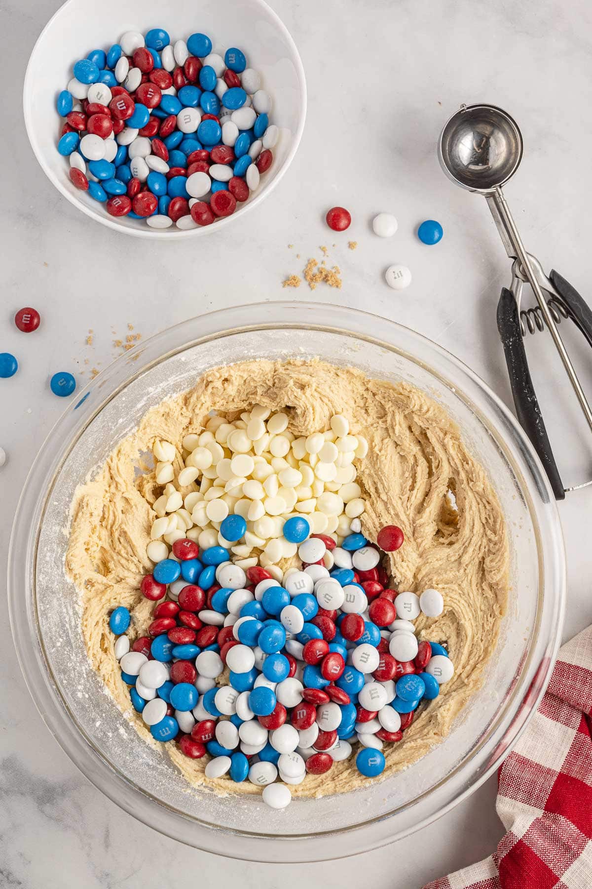 A glass bowl of cookie dough with red, white, and blue candy pieces and white chocolate chips on top, surrounded by a smaller bowl of candy pieces, a scooper, and a red and white cloth on a marble surface.