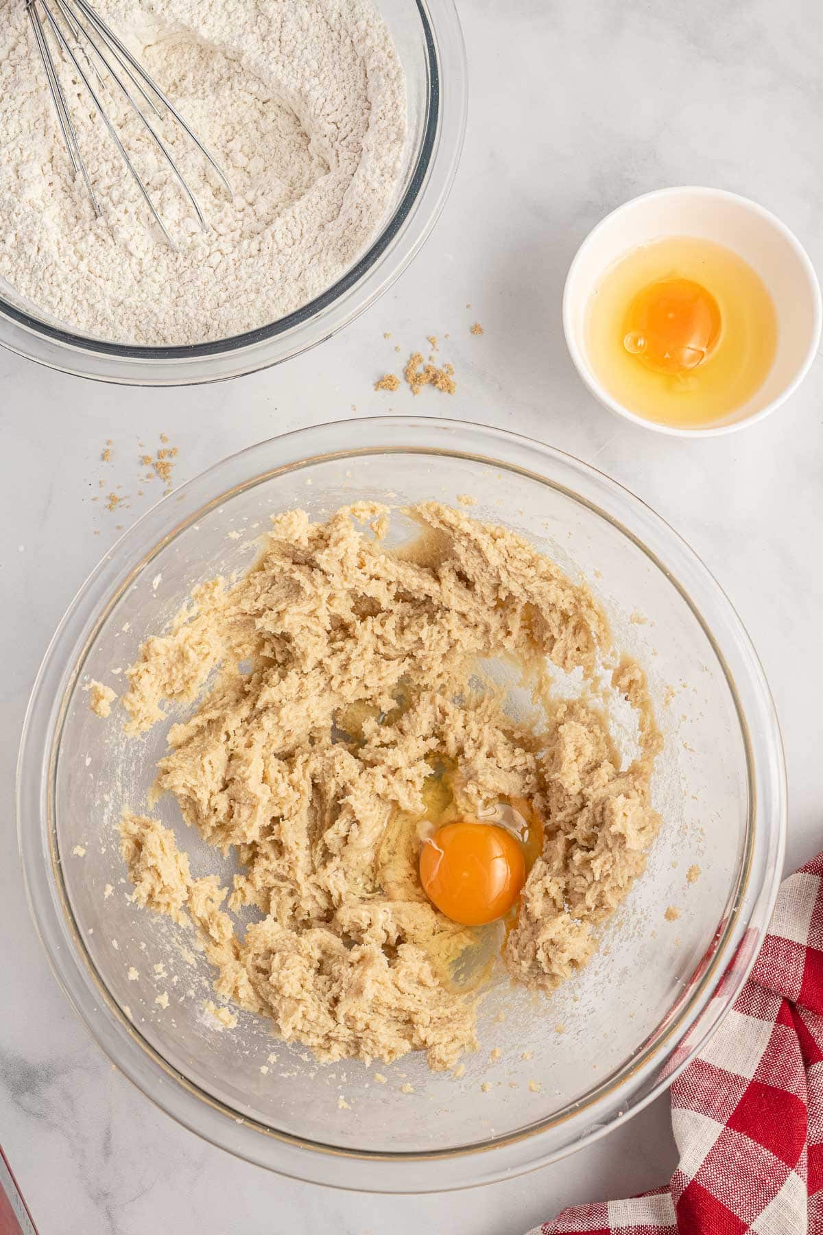 A mixing bowl with cookie dough and an egg, next to a bowl of flour with a whisk, a small bowl containing an egg, and a red checkered cloth on a marble surface.