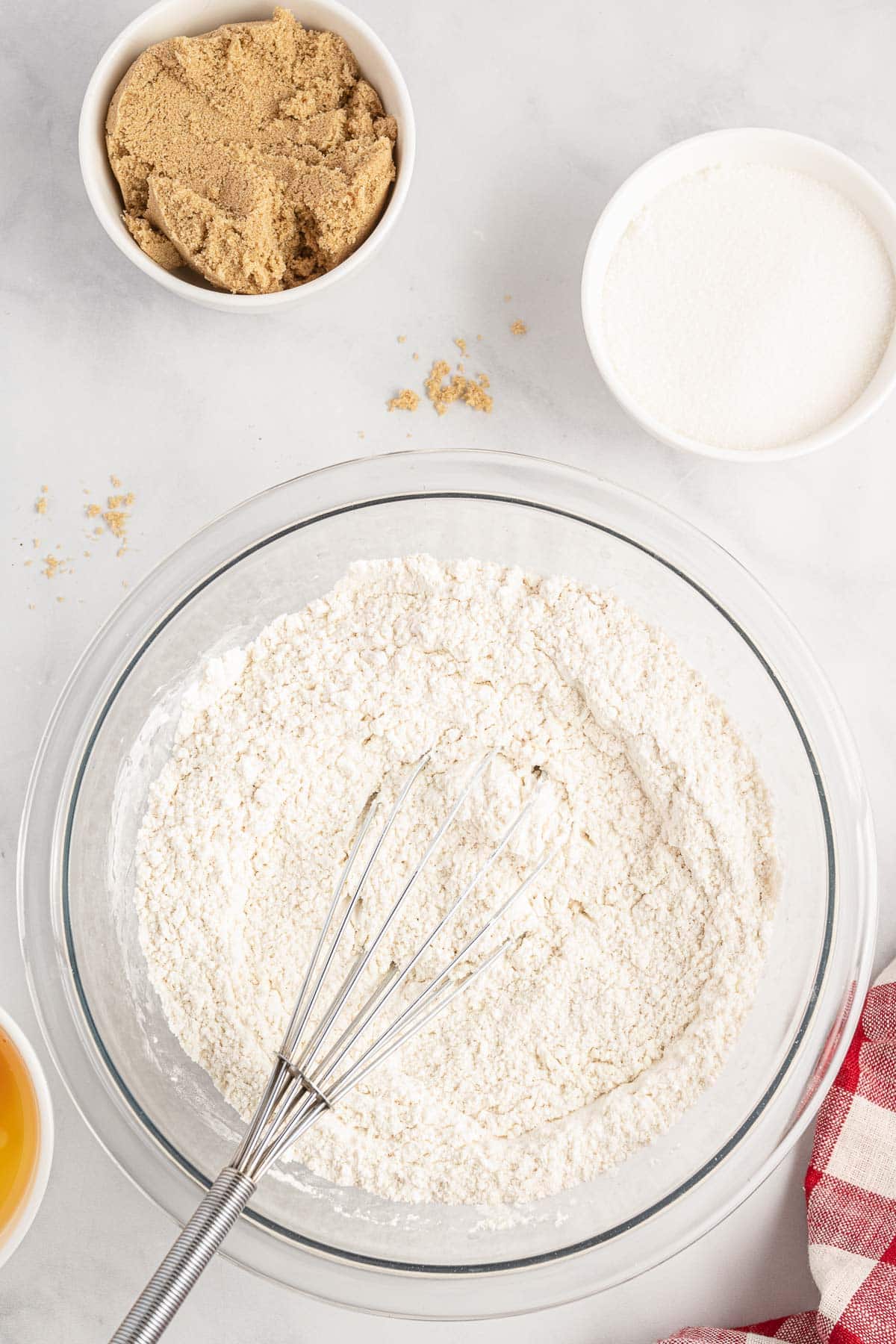 A glass bowl filled with flour and a whisk, surrounded by small bowls containing brown sugar, white sugar, and an egg, all set on a white surface with a red and white checkered cloth nearby.