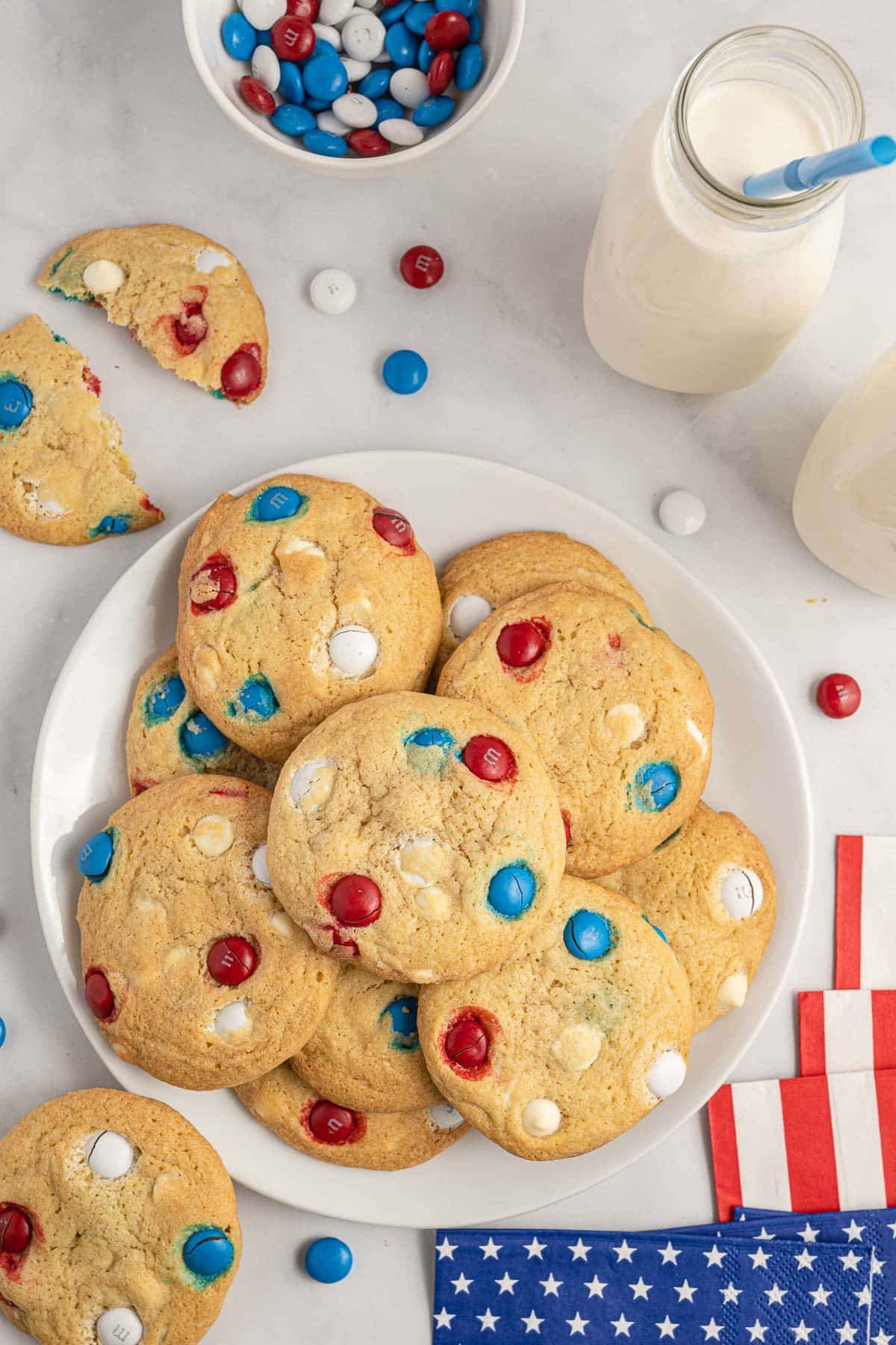 A plate of cookies with red, white, and blue chocolate candies, next to bottles of milk, a bowl of more candies, and American-themed napkins on a white surface.