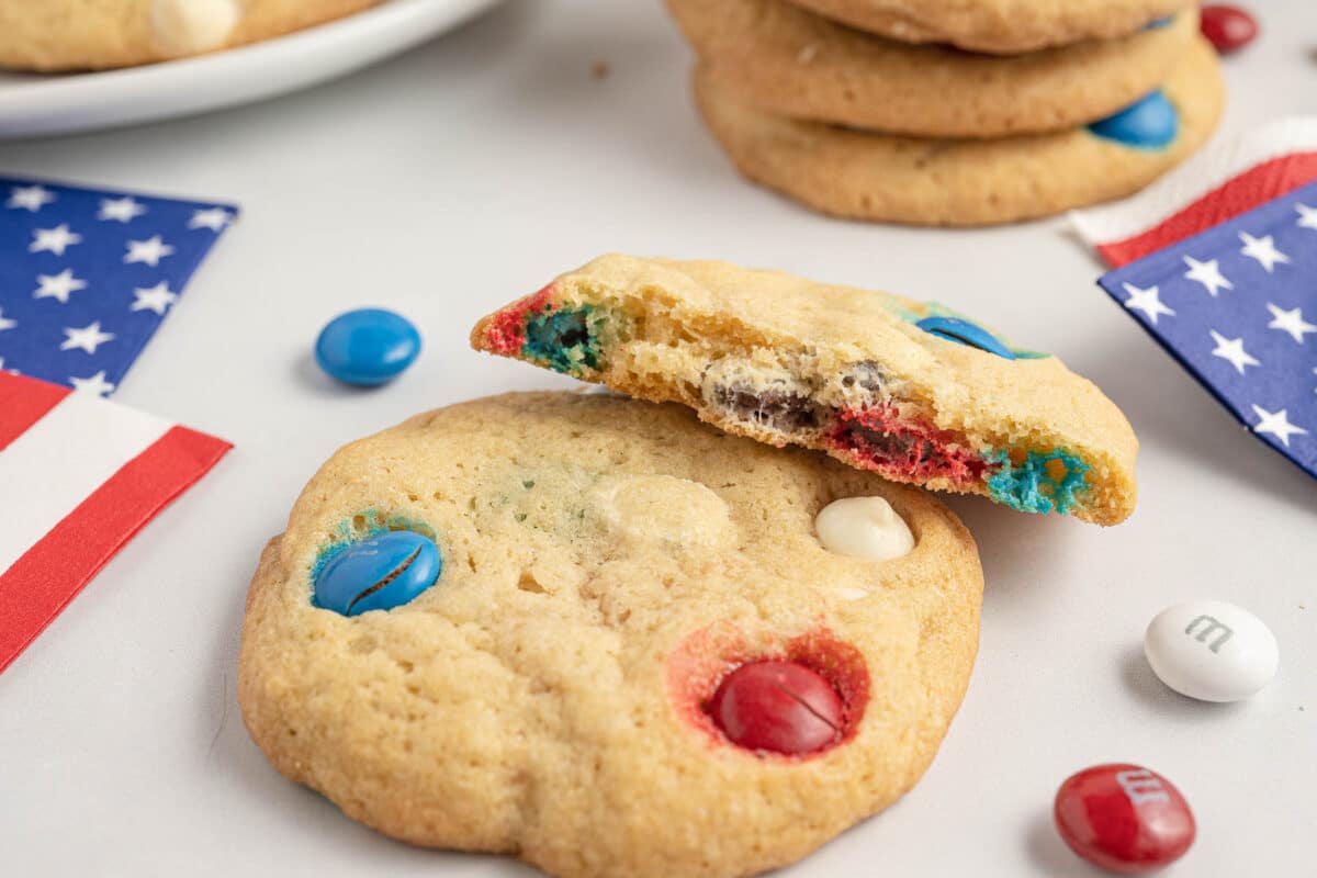 A broken cookie with red, white, and blue candy pieces is in the foreground. Nearby, another cookie, M&M's, and a small American flag are visible on a white surface.