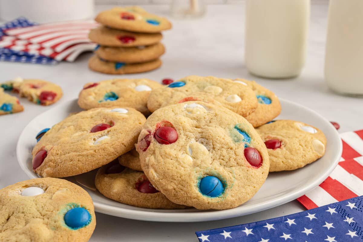 A plate of cookies with red, white, and blue candies on a table decorated with American flag napkins and two glasses of milk in the background.