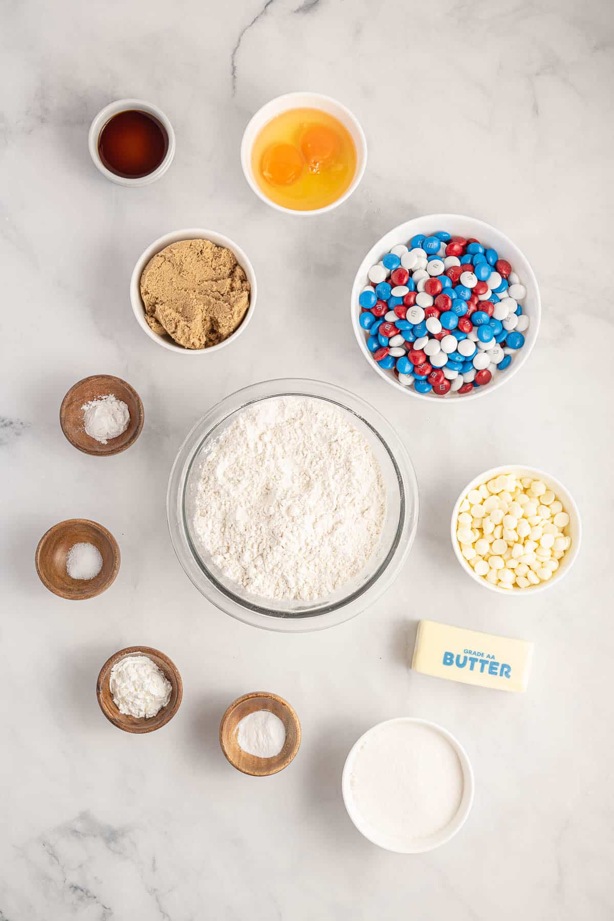 Ingredients for baking cookies arranged on a marble surface, including flour, sugar, butter, brown sugar, vanilla extract, baking soda, baking powder, salt, eggs, chocolate chips, and colorful candies.