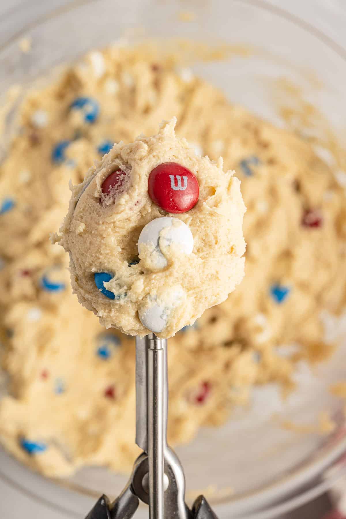 A close-up of a cookie dough scoop with colorful candy pieces against a bowl filled with the same dough in the background.
