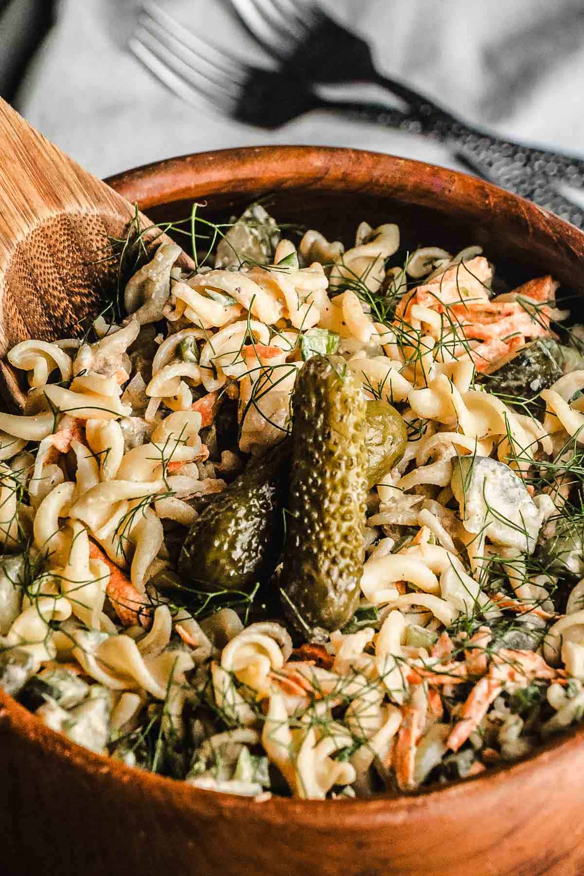 Close-up of a wooden bowl filled with pasta salad, pickles, carrots, and fresh dill, with a wooden spoon.