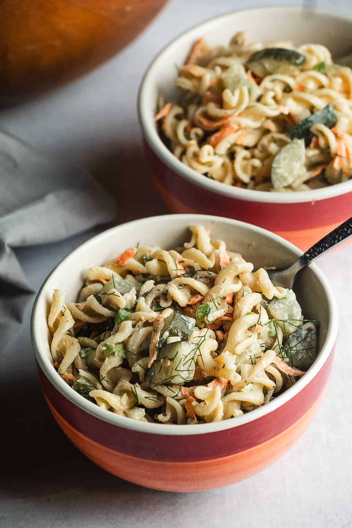 Two bowls of creamy Pickle Pasta Salad with rotini, fresh vegetables, and herbs on a light surface.