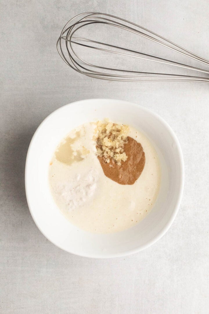 White bowl with creamy liquid, nut butter, grated ginger, and whisk on a light surface—perfect for mixing up your own Pickle Pasta Salad dressing.