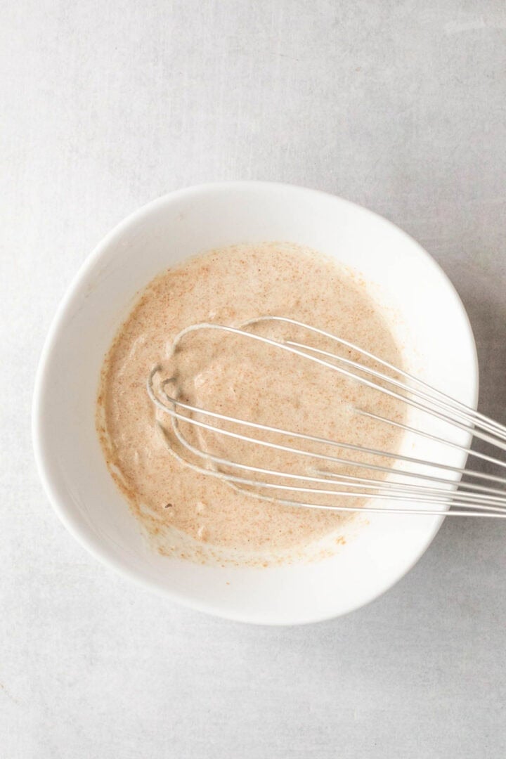 A metal whisk in a white bowl with a light brown batter sits on a light gray surface—perfect for preparing treats to pair with dishes like Pickle Pasta Salad.