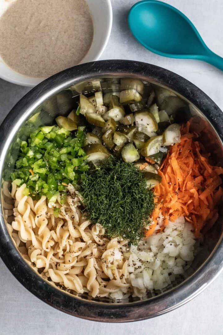 Metal bowl with Pickle Pasta Salad—pasta tossed with chopped pickles, bell pepper, shredded carrot, onion, and dill—served next to a bowl of creamy dressing.