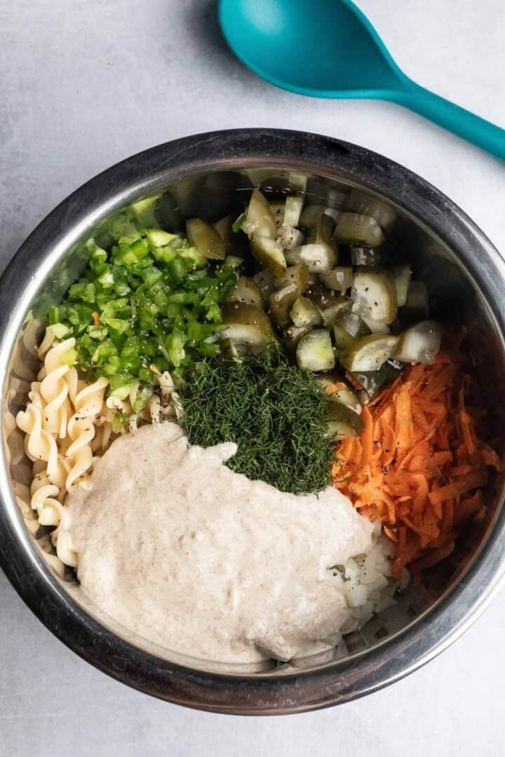 A metal bowl with Pickle Pasta Salad, featuring pasta, chopped veggies, shredded carrot, fresh dill, and a creamy dressing ready to mix.