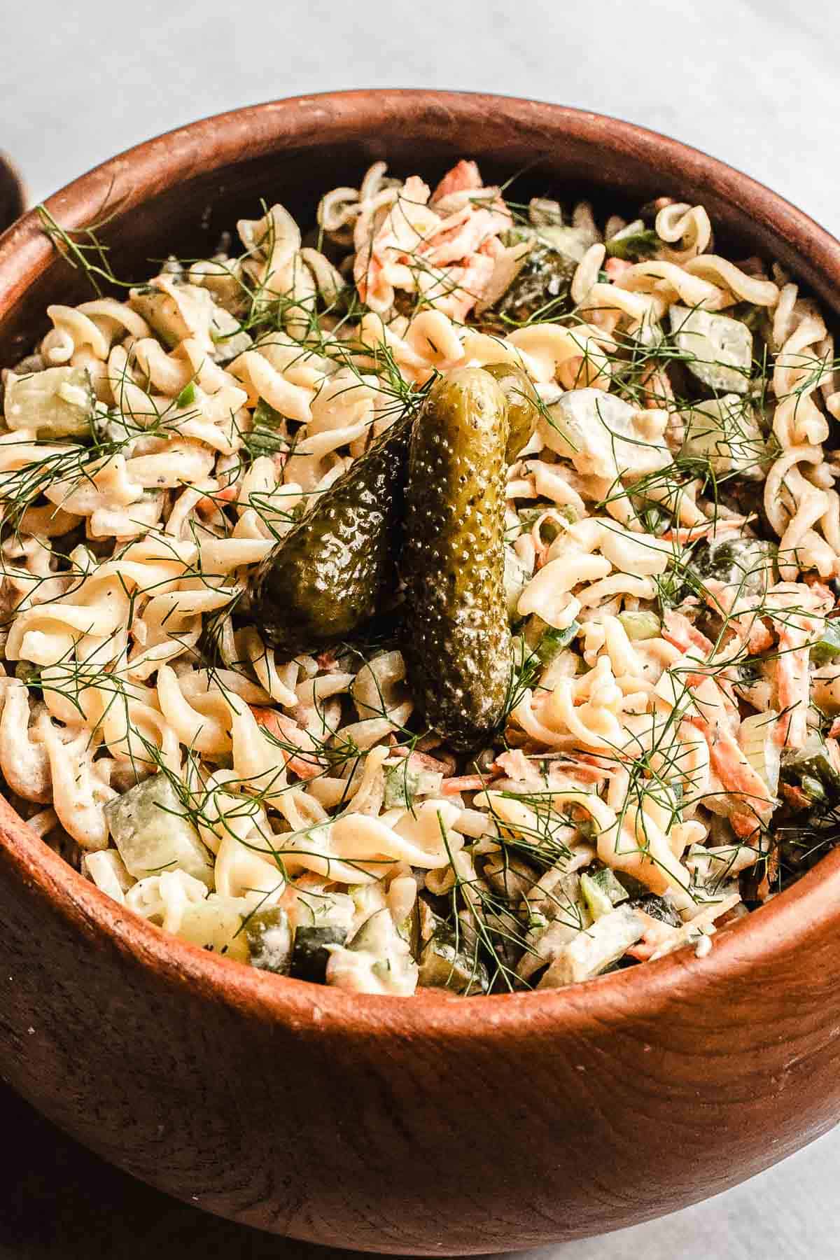 A wooden bowl of creamy Pickle Pasta Salad is topped with two pickles and fresh dill.