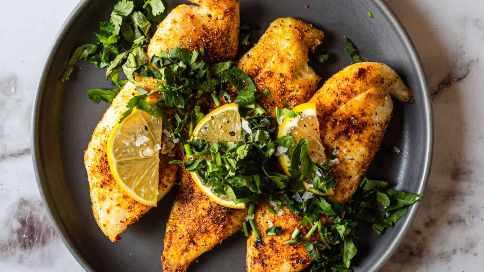 Four seasoned and cooked chicken breasts are arranged on a gray plate, garnished with chopped fresh herbs and lemon wedges. The plate is set on a white marble surface.
