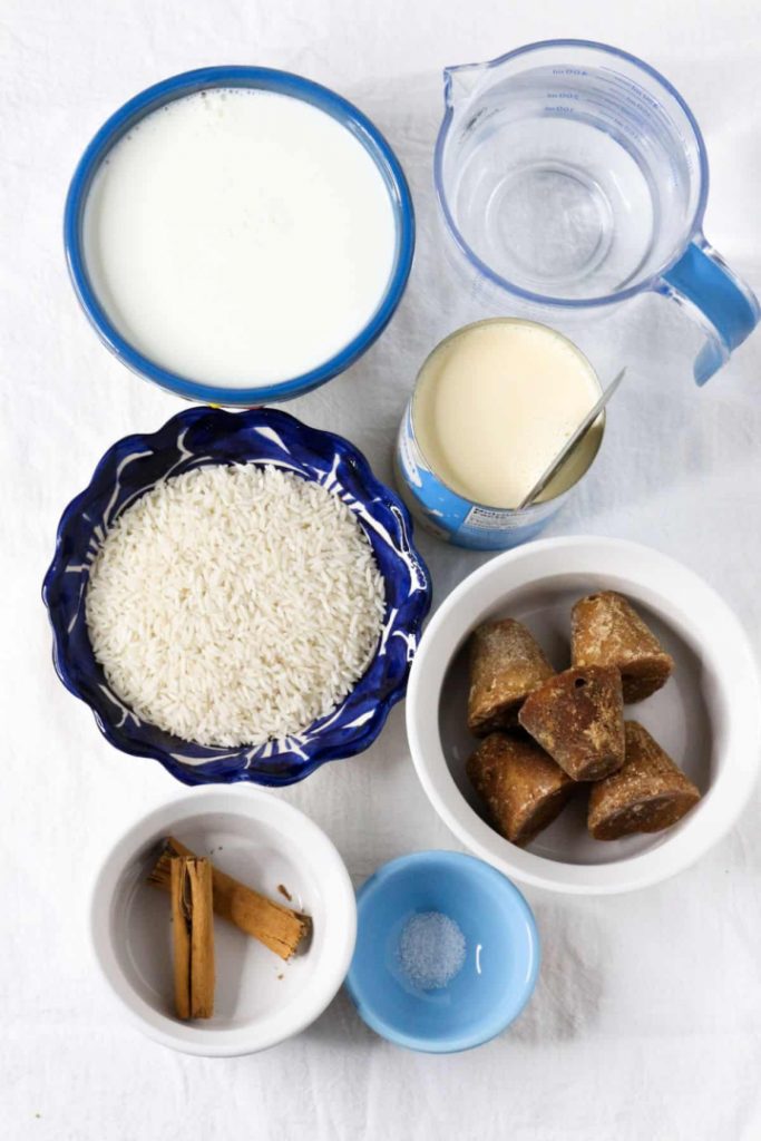 Bowl with milk, measuring cup with water, evaporated milk, white rice, piloncillo cones, cinnamon sticks and salt on a white background