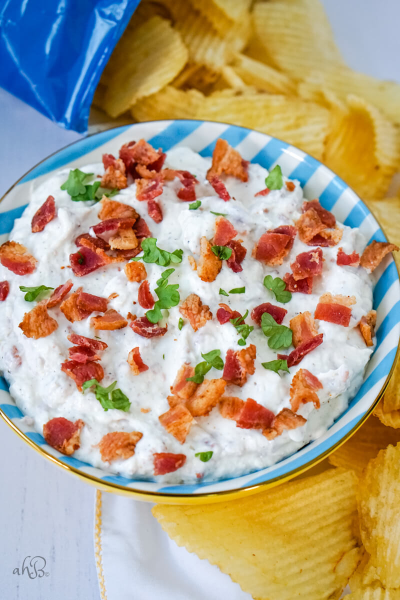 A bowl of bacon horseradish dip is served in a blue and white striped bowl and surrounded by potato chips.