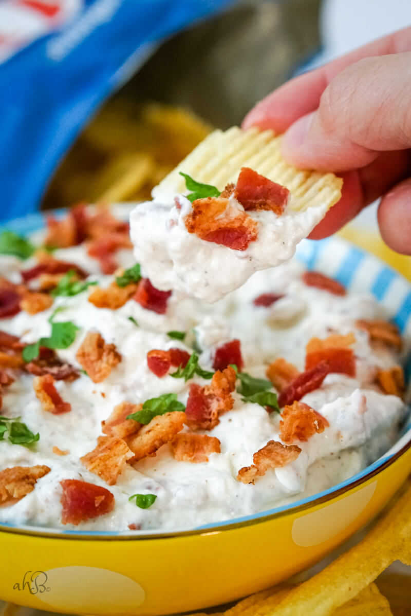 A chip scoops up bacon horseradish dip from a blue and white striped bowl.