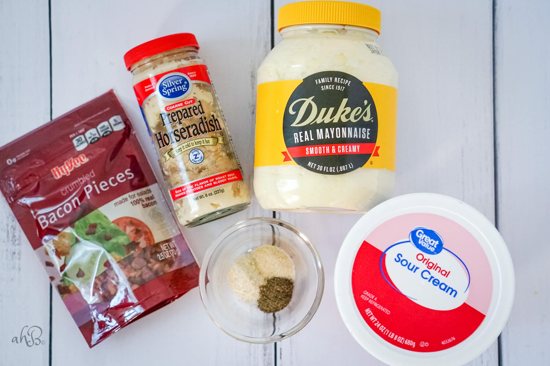 A package of bacon pieces, prepared horseradish, a dish of seasonings, real mayonnaise, and sour cream sit on a table.