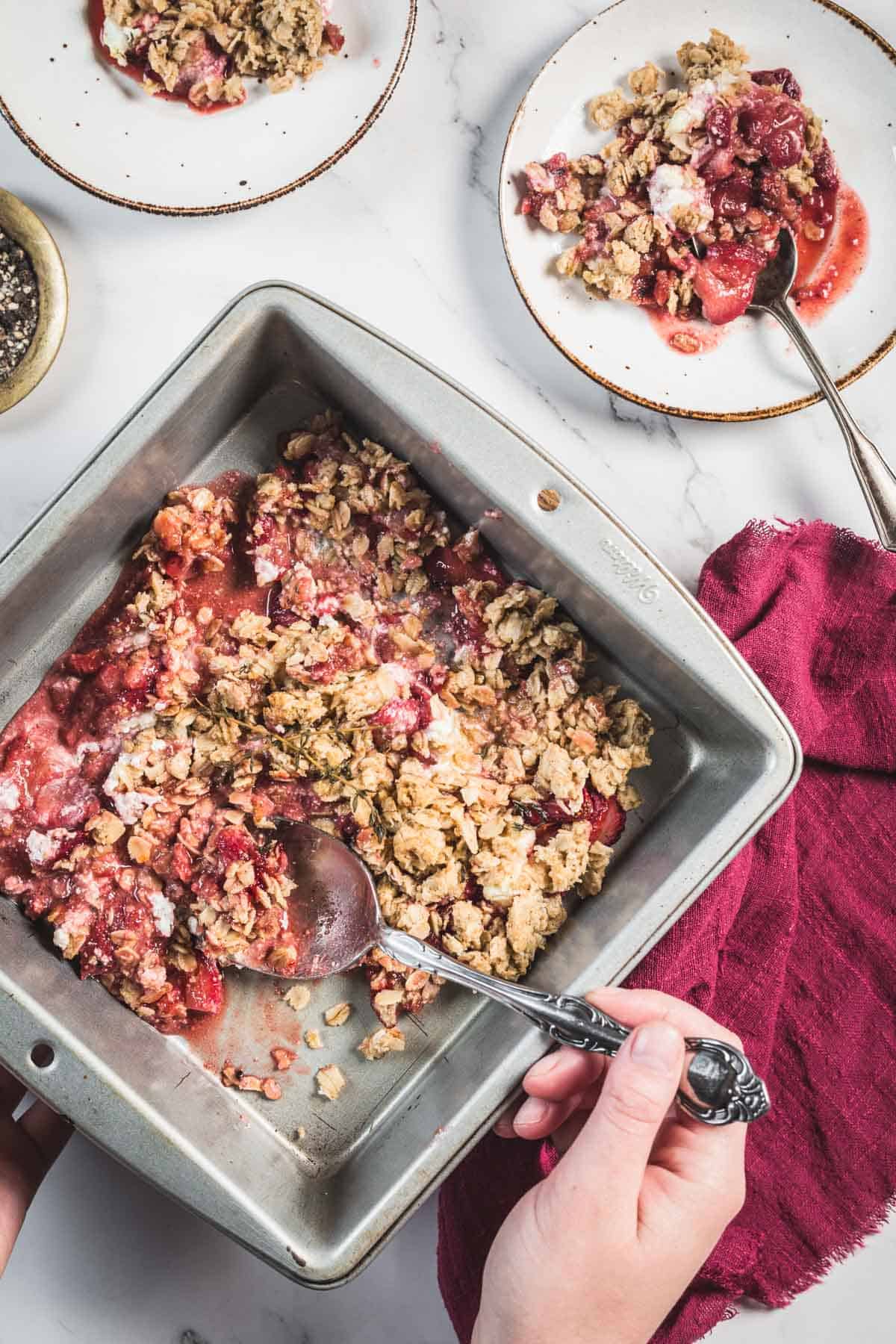 A hand holds a metal baking dish containing a partially served Balsamic Strawberry Crisp with a spoon. Two plates with portions of the crisp and spoons are visible on a white marble surface, along with a red cloth napkin.