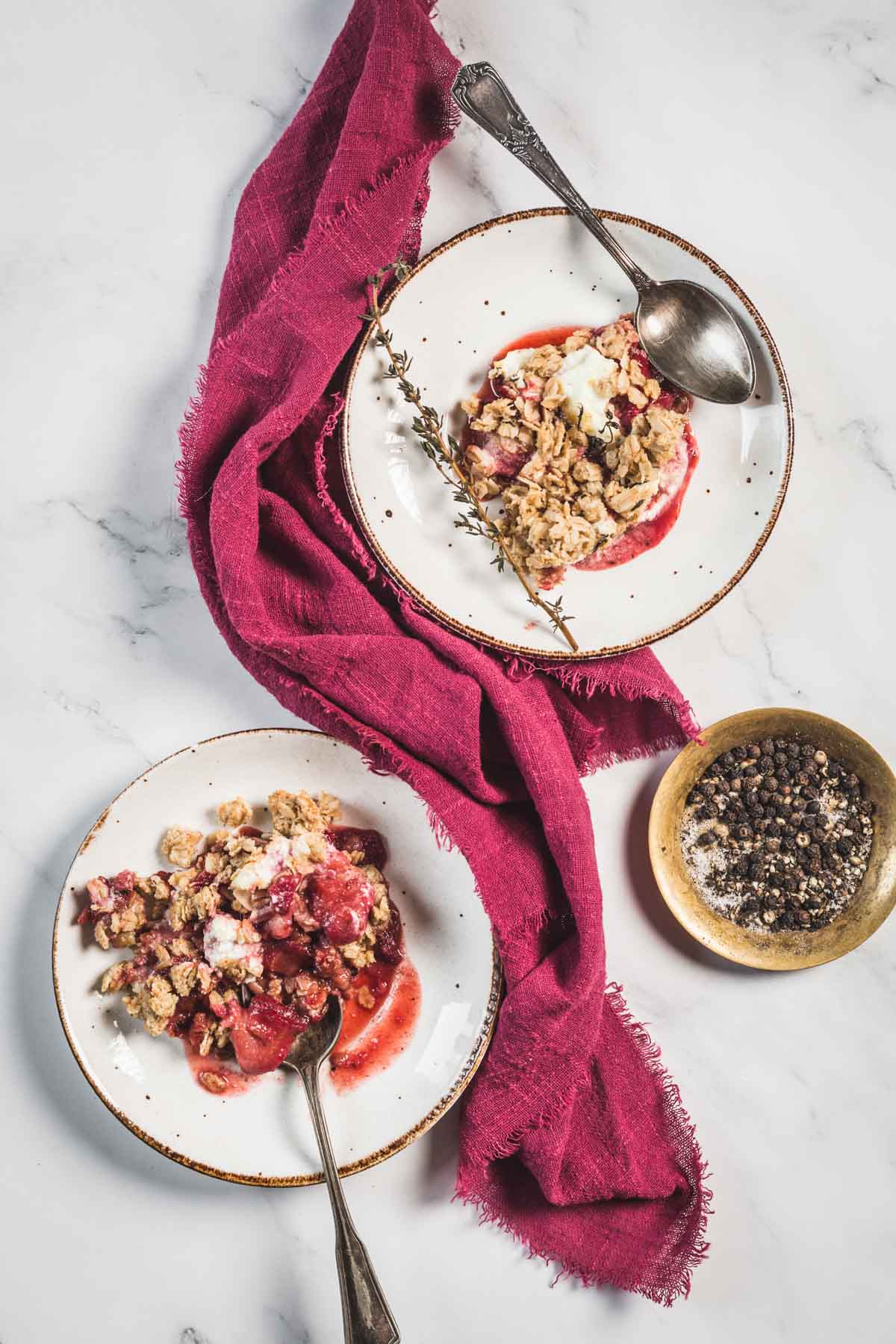 Two plates with servings of Balsamic Strawberry Crisp and whipped cream sit on a marble surface, each with a spoon. A burgundy napkin runs between them, and a small bowl of peppercorns sits nearby.