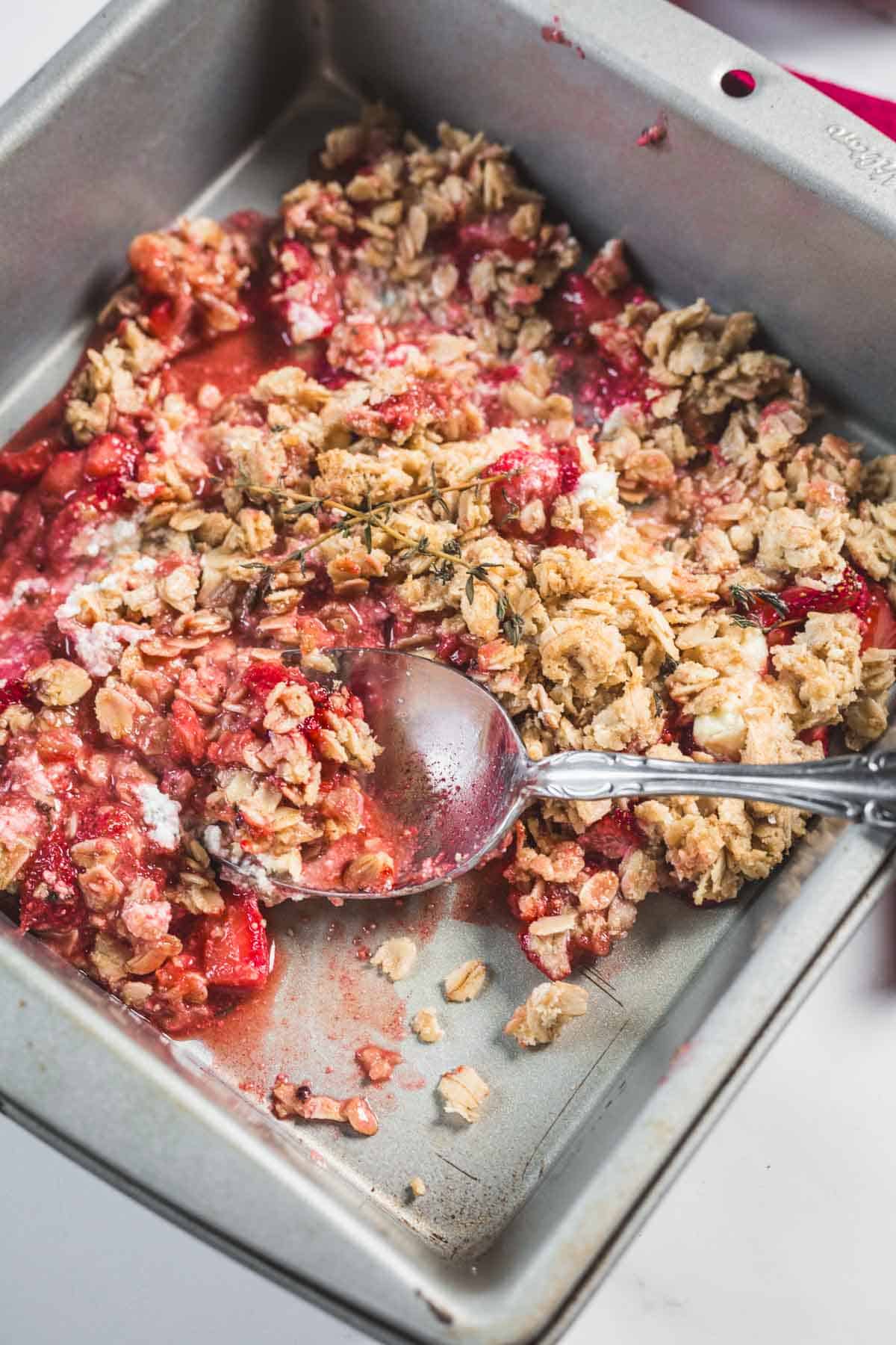 A square metal baking pan containing a partially served Balsamic Strawberry Crisp with a crumbly oat topping and red fruit filling. A metal spoon rests in the dish, with some filling and crumbs on it.