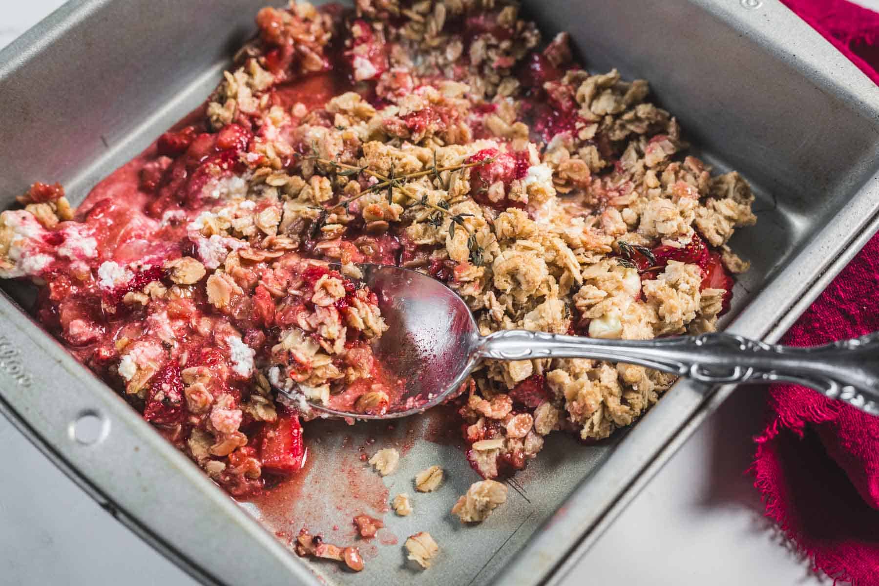 A metal baking pan containing a partially served Balsamic Strawberry Crisp with an oat topping and a silver spoon. Some of the baked fruit and oat crumble mixture has been scooped out, revealing the red strawberry balsamic dessert filling.