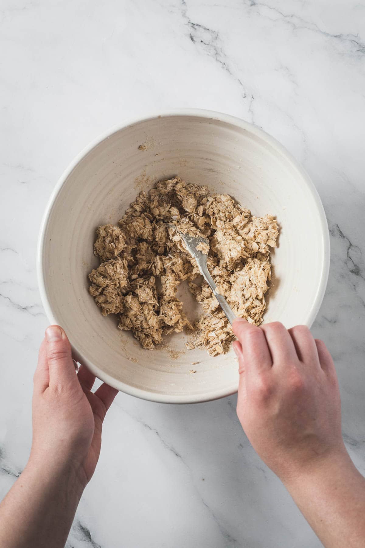 Two hands hold a white bowl and mix lumpy dough for a Balsamic Strawberry Crisp with a metal utensil. The bowl rests on a white marble surface.