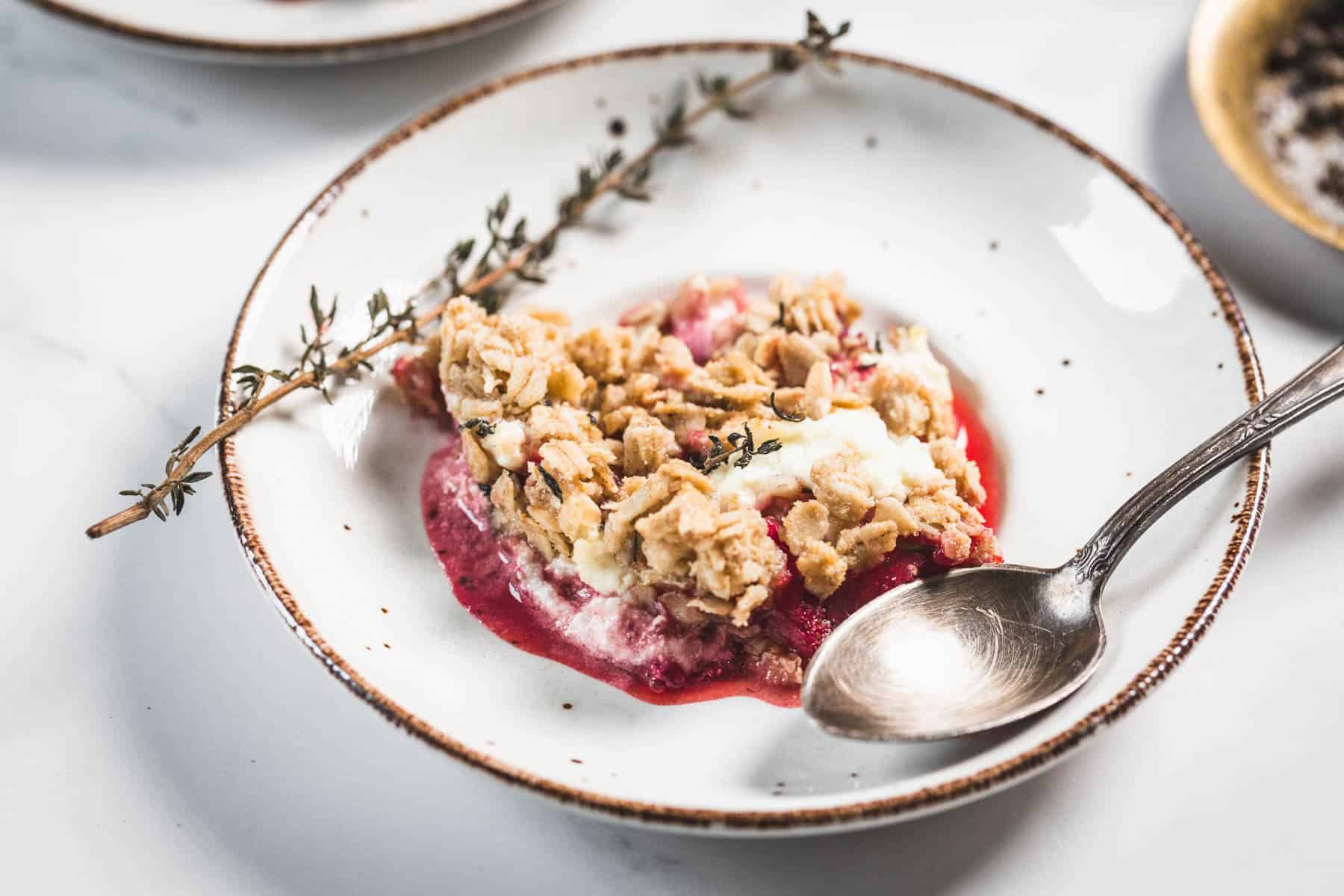 A white plate with a serving of balsamic strawberry crisp topped with oat streusel, some melted ice cream, a sprig of herbs, and a metal spoon resting on the side.
