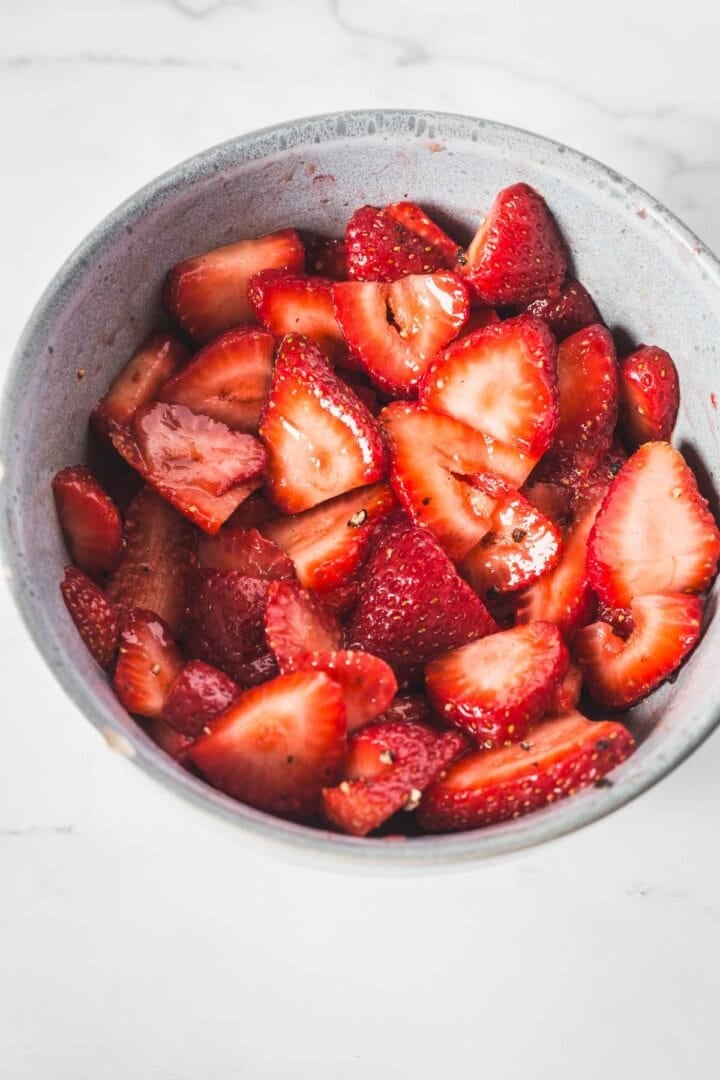 A gray bowl filled with sliced strawberries sits on a white marble surface, perfect for preparing a fresh Balsamic Strawberry Crisp. The strawberries appear vibrant, fresh, and slightly glossy.