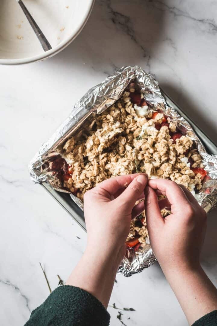 A person is sprinkling a crumbly topping over a foil-lined pan of Balsamic Strawberry Crisp. A mixing bowl with a spoon sits on the white marble countertop, setting the scene for a delicious strawberry dessert.