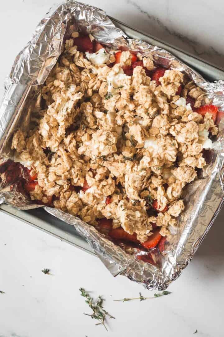 A baking pan lined with foil is filled with sliced strawberries, topped with a crumbly oat mixture for a delicious Strawberry Crisp, sitting on a light-colored countertop. Some fresh herb sprigs are scattered nearby.
