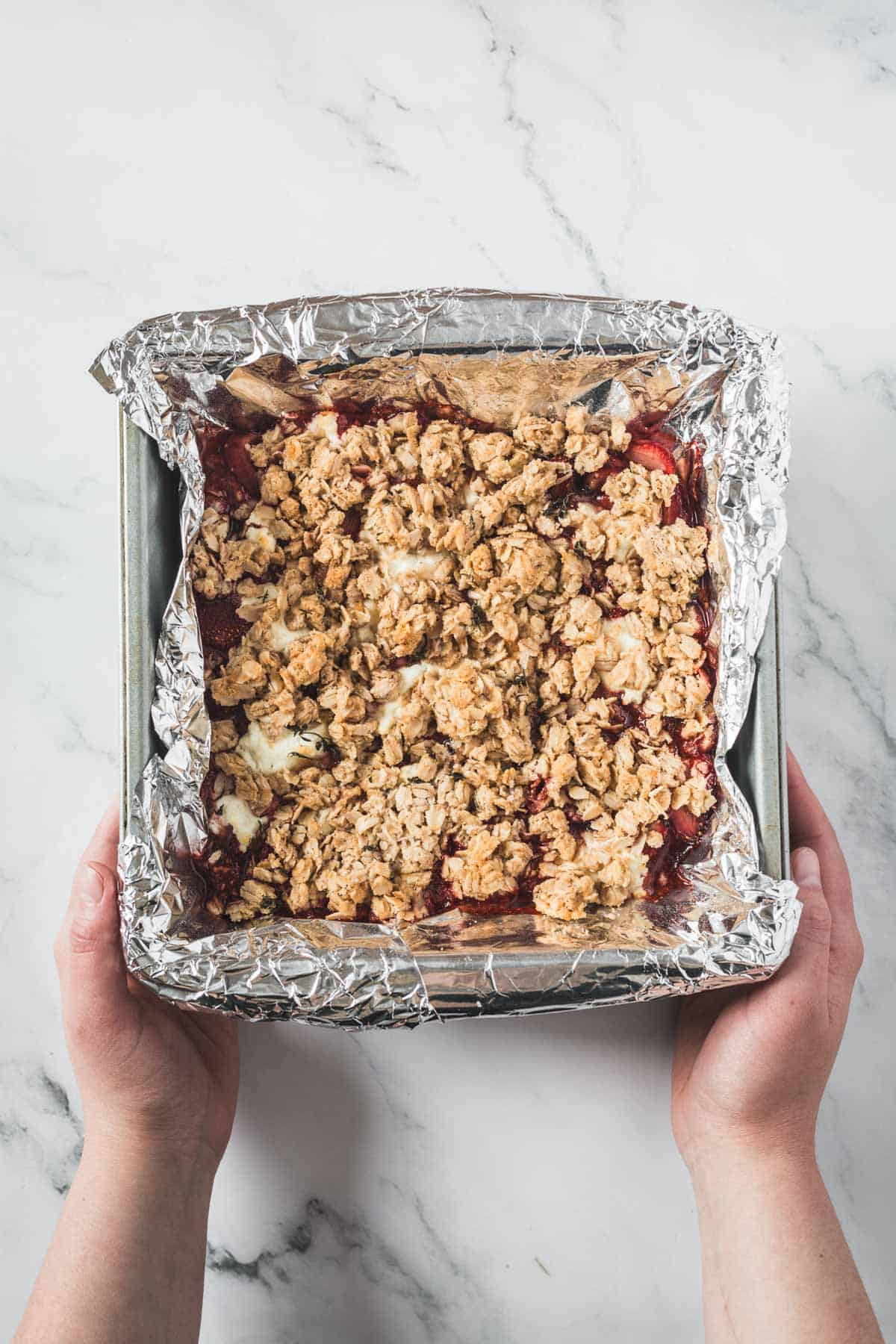 A pair of hands holds a square baking pan lined with aluminum foil and filled with a freshly baked Balsamic Strawberry Crisp, resting on a white marble surface.