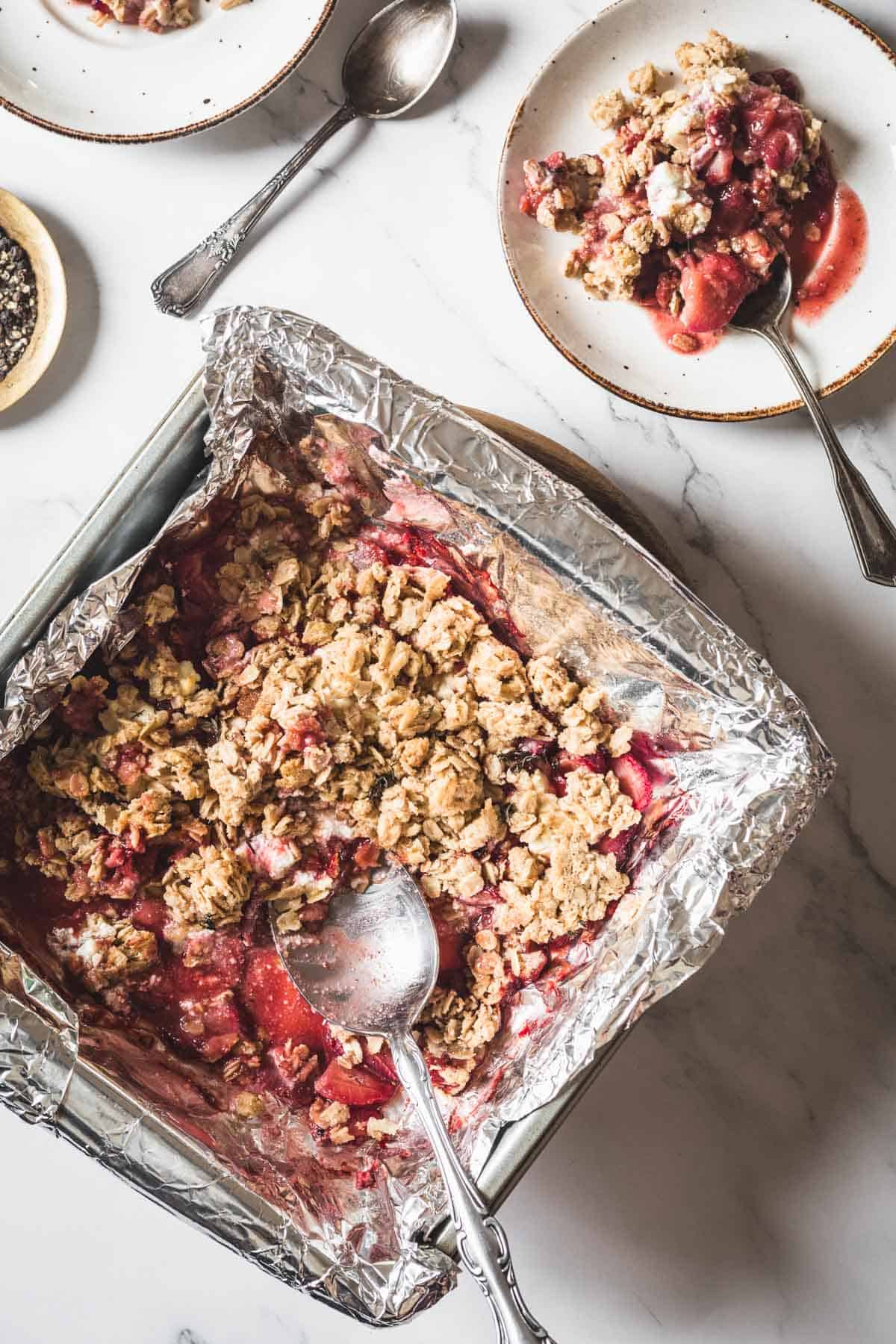A square baking dish lined with aluminum foil holds a Balsamic Strawberry Crisp and oat crumble dessert. A large spoon rests in the dish, while two plates with servings of the crisp and spoons are nearby on a marble surface.