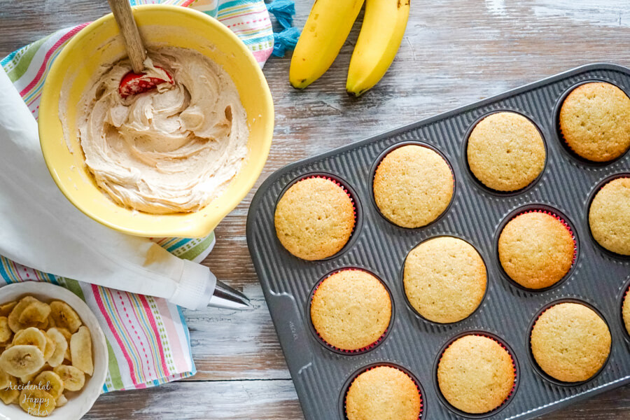 A bowl of peanut butter frosting and a bowl of banana chips next to a pan of cupcakes ready to be frosted and decorated. 