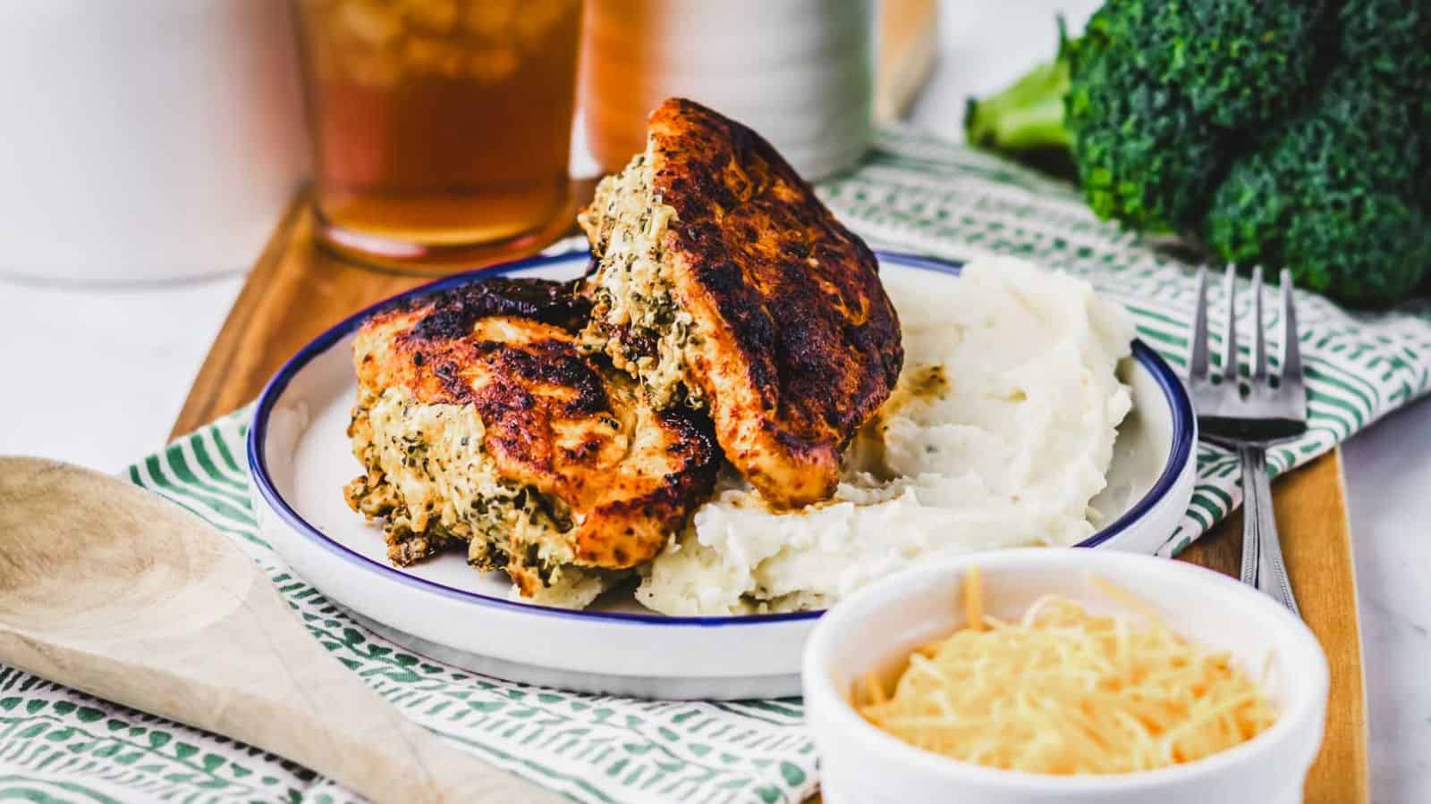 Two pieces of stuffed and grilled chicken breast with a browned, crispy exterior are served on a plate next to a portion of mashed potatoes. The chicken stuffing appears to contain herbs and cheese.