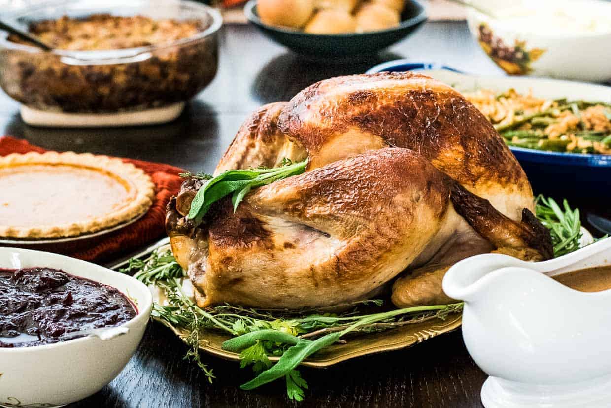 A roasted turkey garnished with herbs is placed at the center of a dining table surrounded by various dishes, including cranberry sauce, pie, green beans, stuffing, and a gravy boat—a feast filled with Thanksgiving-worthy recipes to wow everyone.