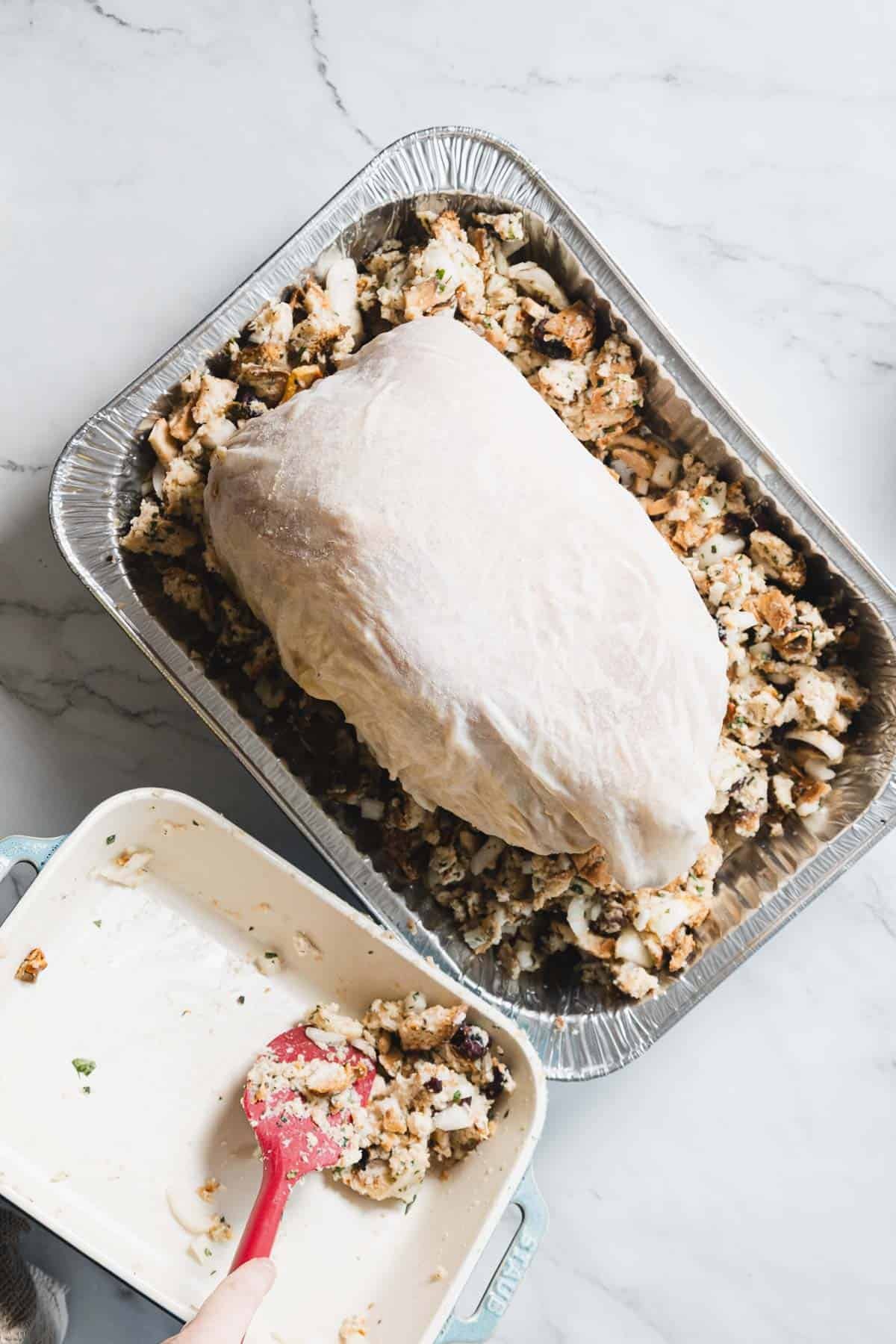 A raw, butterflied turkey breast sits on top of bread stuffing in a large aluminum tray, with extra stuffing being added from a smaller dish using a red spatula. Cheesecloth is ready nearby for wrapping before roasting on the marble countertop.