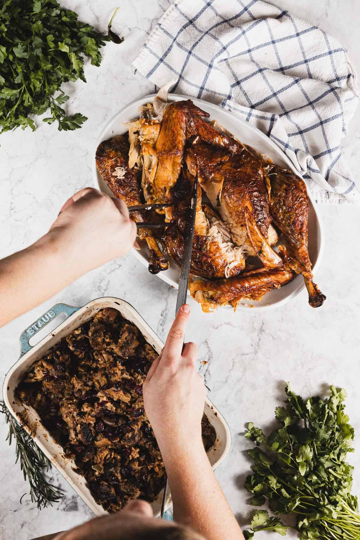 A person carves a roasted turkey, perfectly golden from roasting with cheesecloth, on a white platter. Beside it is a dish of stuffing. Fresh parsley and a blue checkered towel are on the marble countertop.