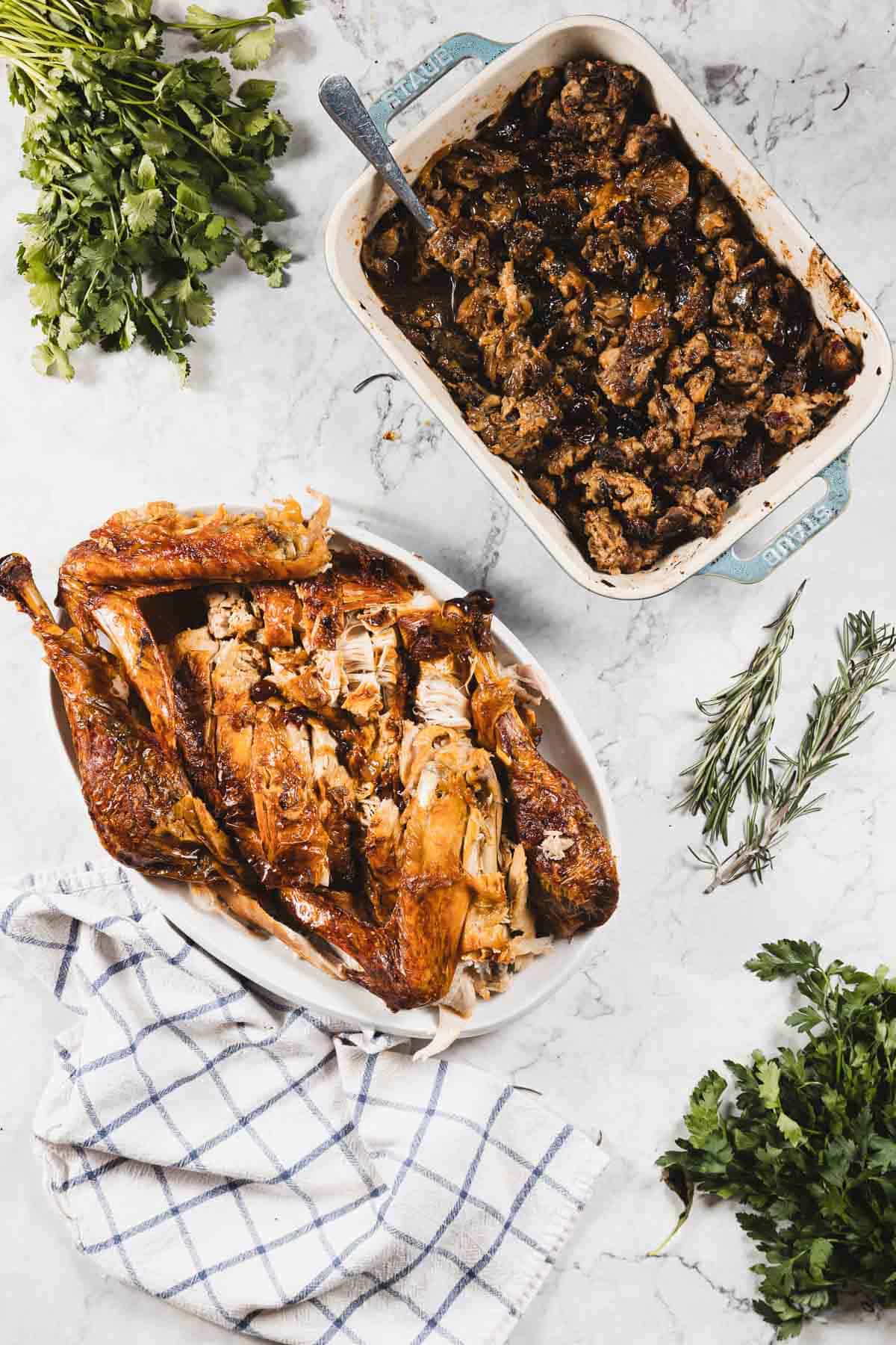 A serving dish with roasted, carved turkey, a baking dish of browned stuffing, fresh herbs, and a checkered cloth on a white marble surface. The golden skin hints at careful roasting with cheesecloth for extra moisture.