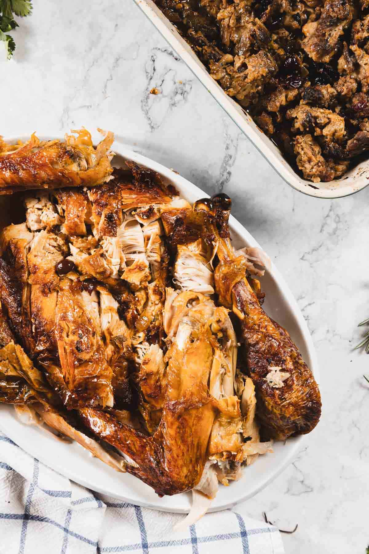 A serving dish filled with carved roasted turkey pieces, cooked to perfection using cheesecloth, sits on a marble surface next to a baking dish of stuffing. A checkered cloth and fresh herbs complete the inviting scene.