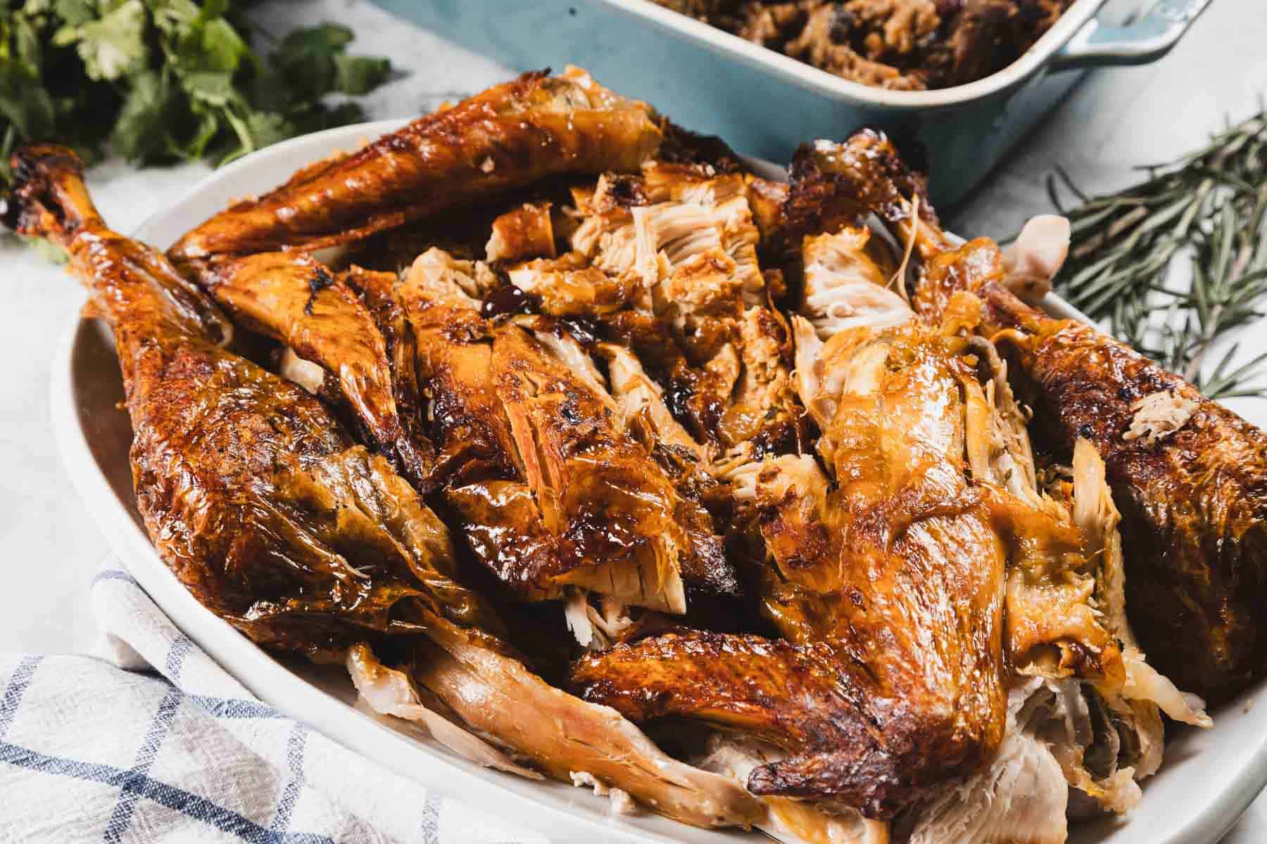 A white serving dish filled with pieces of roasted turkey, its golden-brown crispy skin perfected with cheesecloth, sits on a white surface with herbs and a blue baking dish in the background.