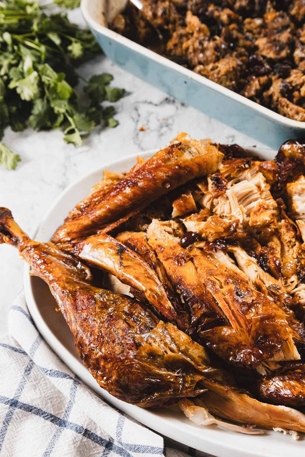 A platter of carved roasted turkey with golden-brown skin—roasted to perfection with cheesecloth—sits on a white dish next to a blue towel, with a pan of stuffing and fresh parsley in the background.
