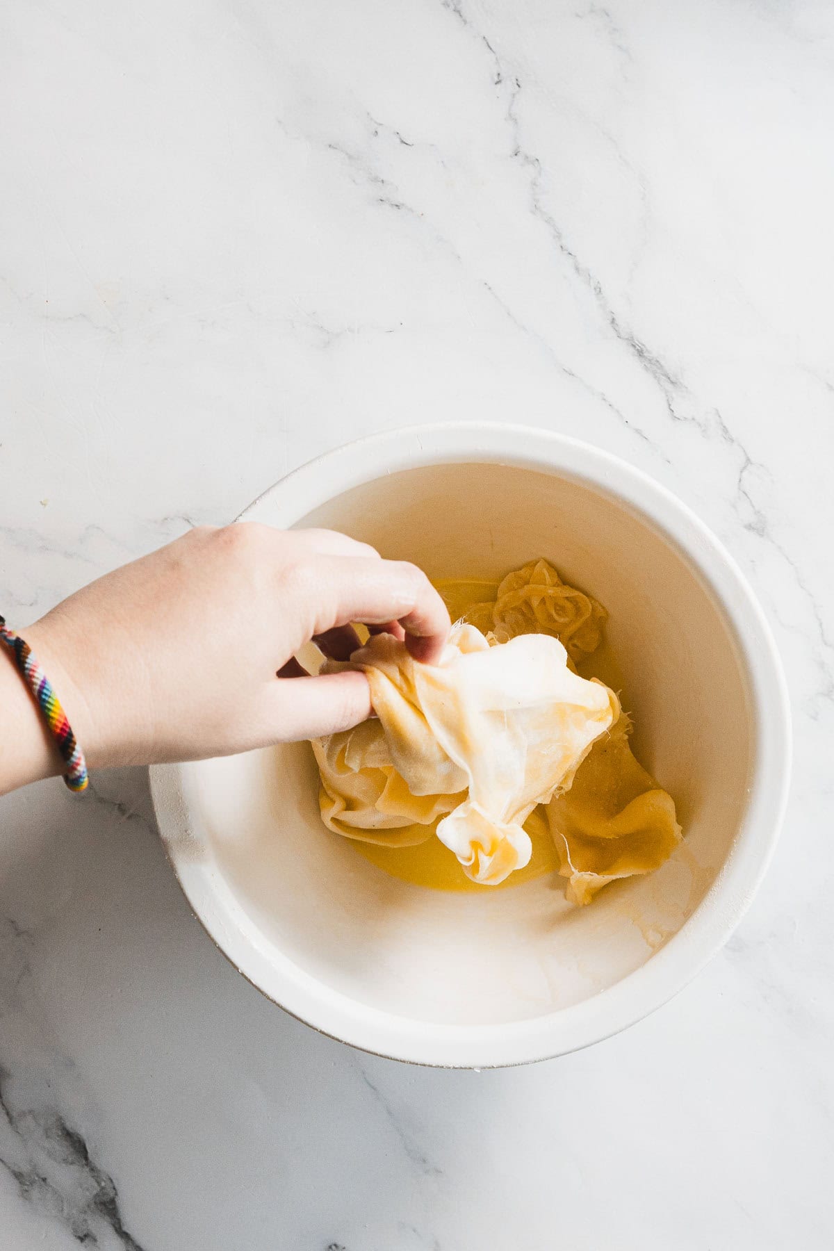 A hand holding a kombucha SCOBY above a white bowl filled with liquid, on a white marble surface—ideal for straining like turkey through cheesecloth.