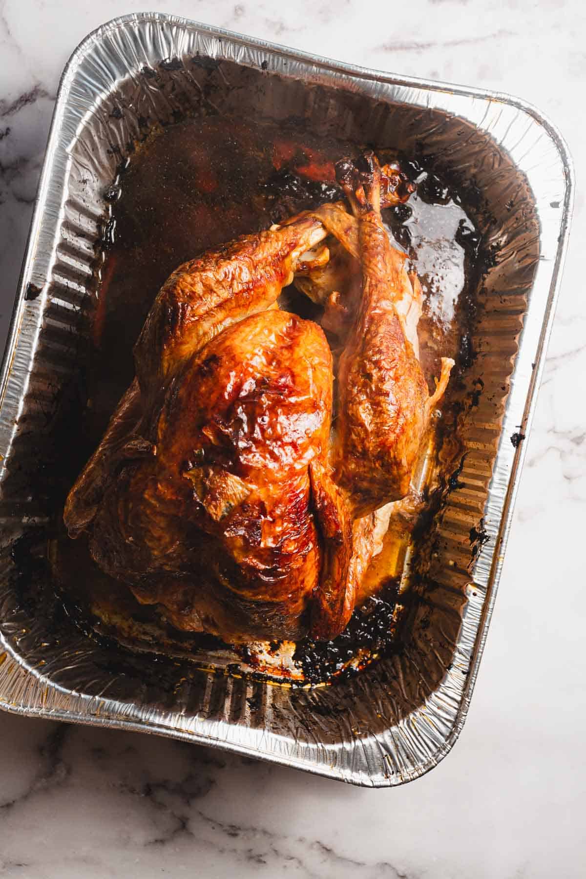 A whole roasted turkey covered with cheesecloth boasts browned skin in a disposable aluminum roasting pan. Juices collect at the bottom, and a marble surface is visible underneath.