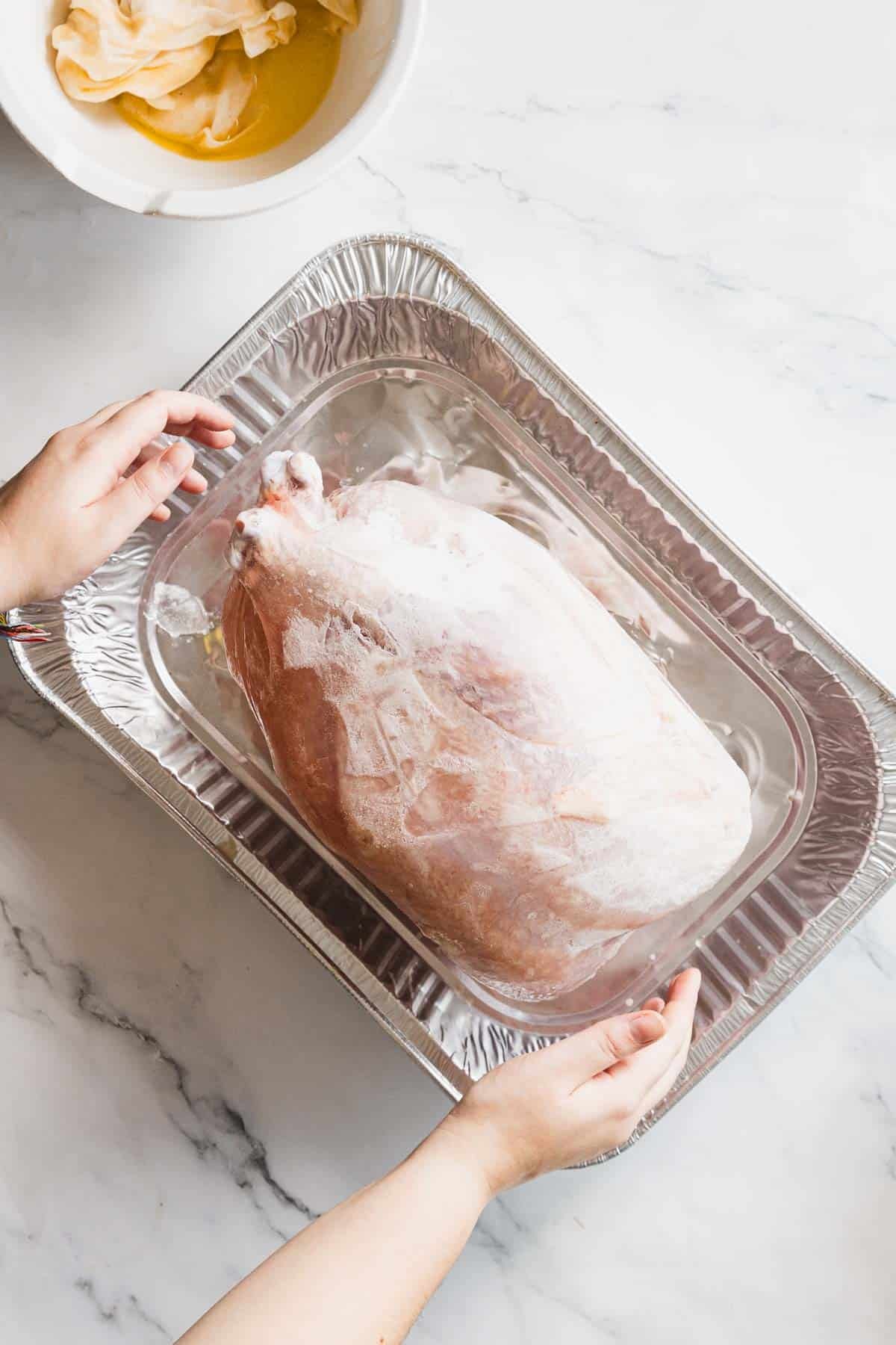 A person holds a large frozen turkey breast submerged in water inside a disposable aluminum roasting pan; in the upper left corner, a bowl with yellow liquid and cheesecloth is visible.