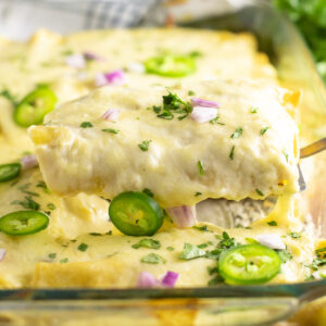 Two cheesy chicken enchiladas being lifted from a casserole dish filled with enchiladas topped with cheese sauce, fresh cilantro, jalapeno slices, and chopped red onion.
