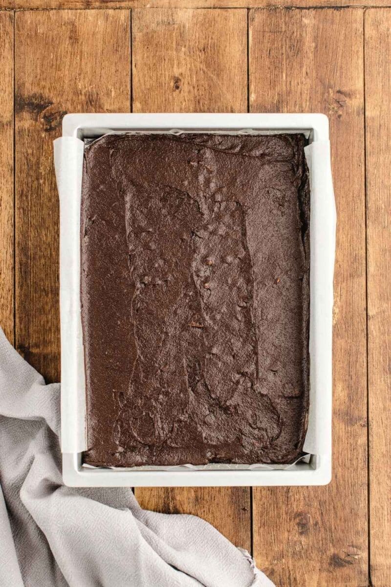A square baking pan filled with unbaked chocolate brownie batter, lined with parchment paper, sits on a wooden surface next to a light gray cloth.
