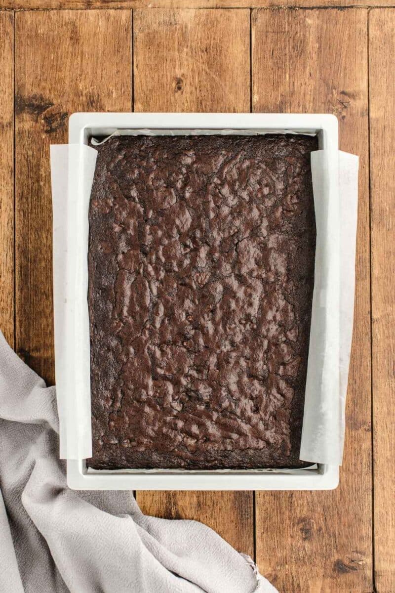 A rectangular pan lined with parchment paper contains a batch of baked brownies on a wooden surface next to a light gray cloth.