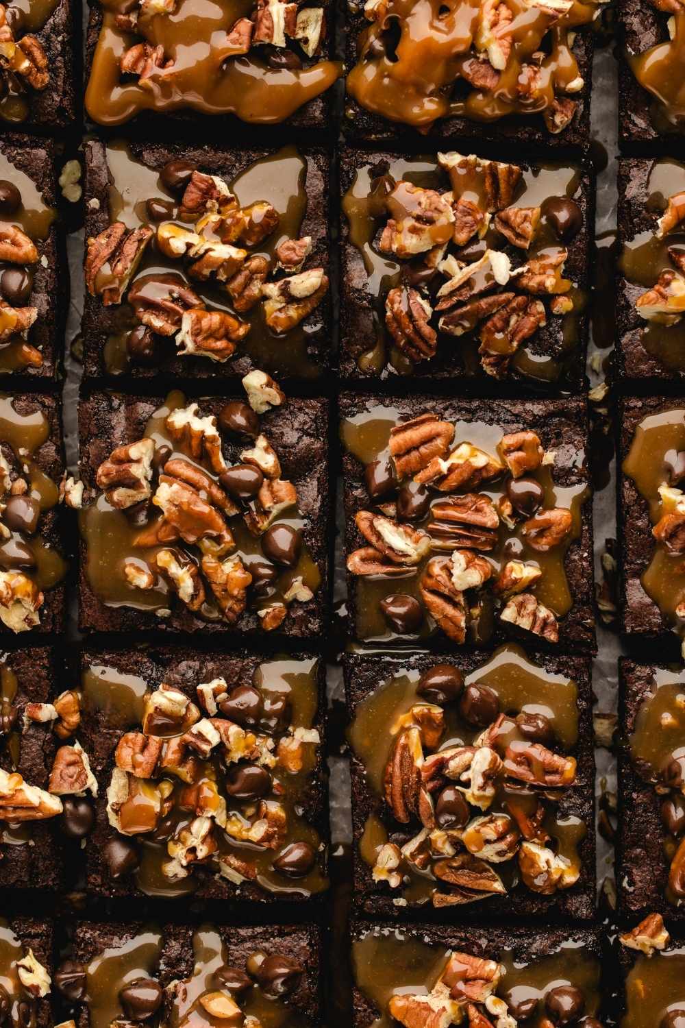 Close-up of several chocolate brownies topped with pecans, caramel sauce, and chocolate chips, arranged in a grid pattern.