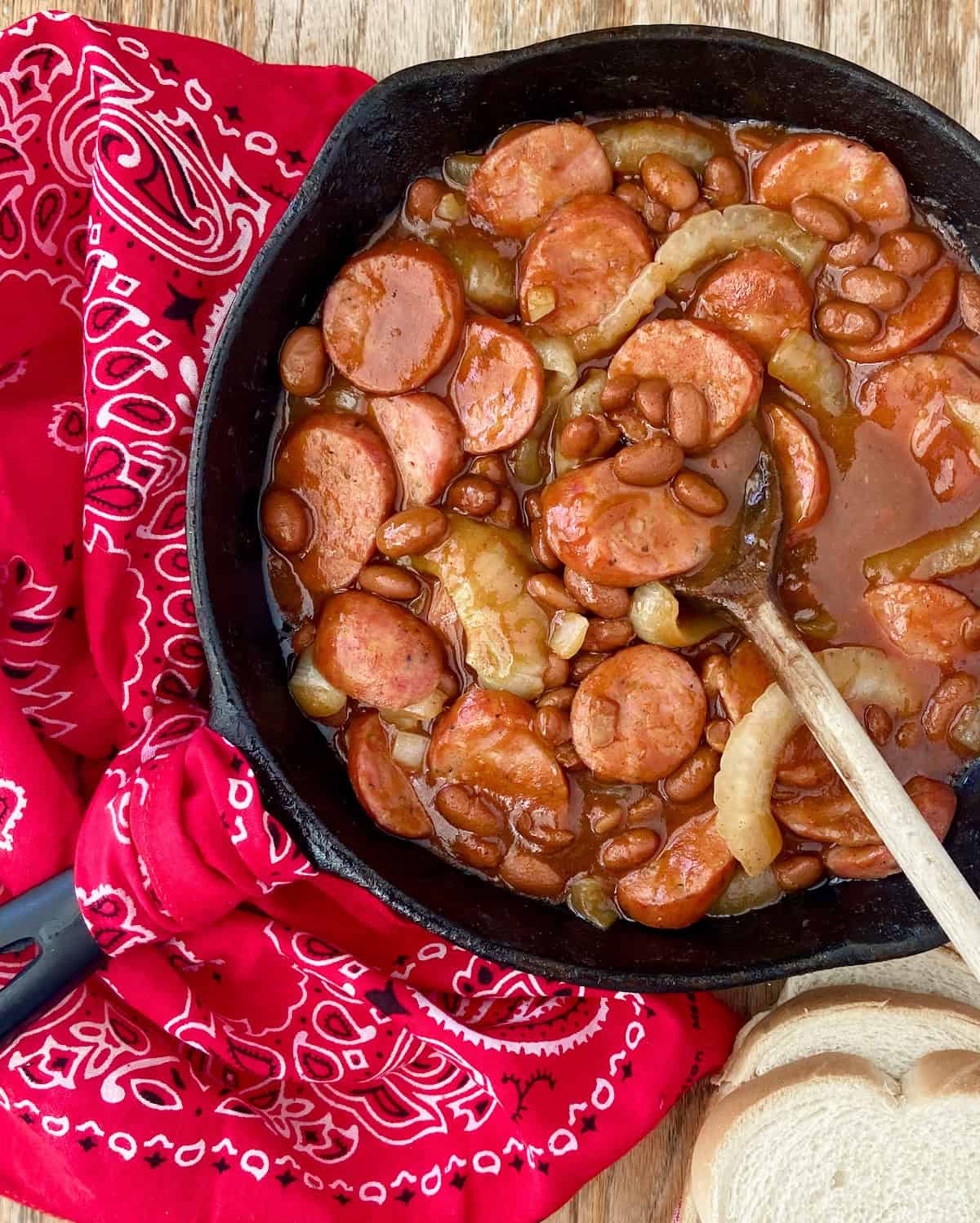 Beans, sausage and onions in cast iron skillet with wooden spoon.